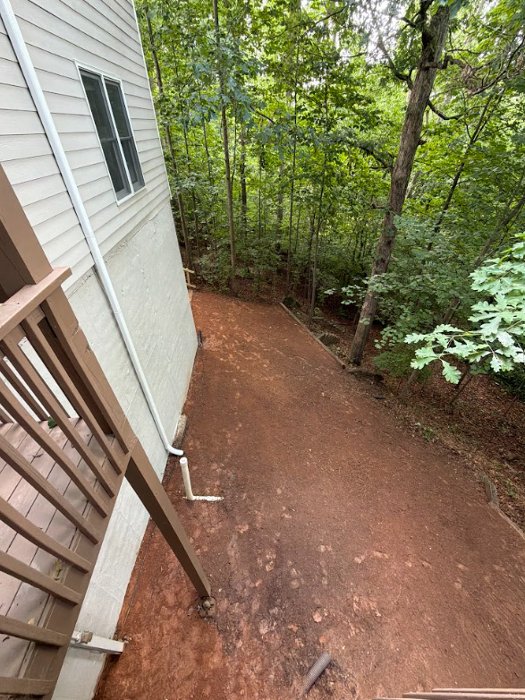 Side yard cleared of brush and graded to bare soil beside a home with the tree line left standing