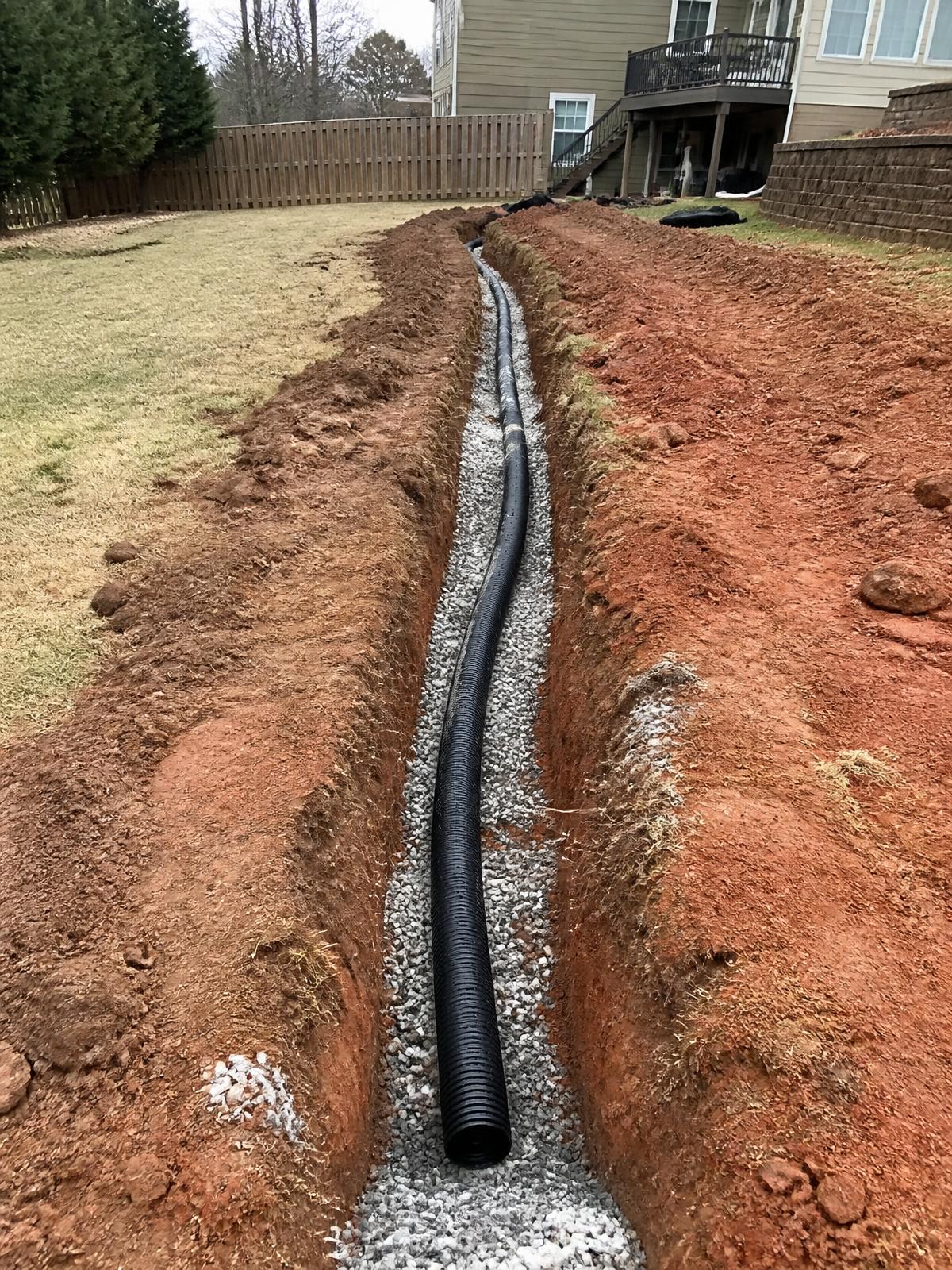 Long drainage trench with perforated pipe bedded in gravel running across a backyard toward the house