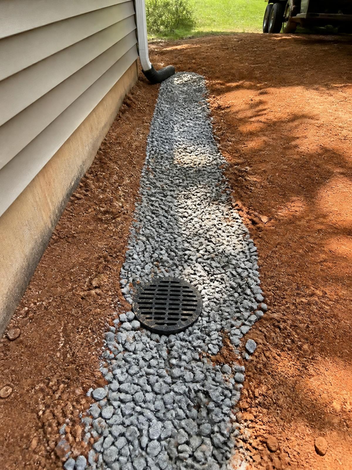 Finished French drain with a round catch basin set in gravel along the side of a house, tied into the downspout