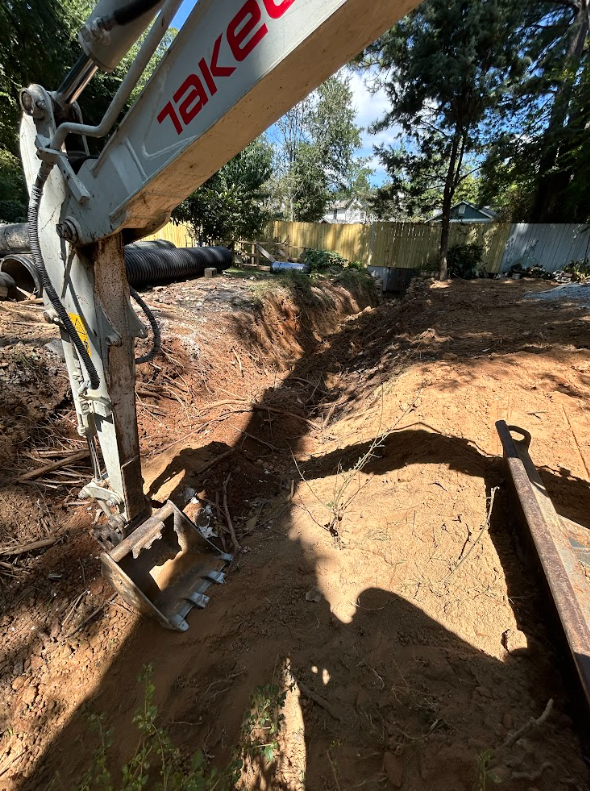 Compact excavator digging a deep trench across a residential backyard with drainage pipe staged nearby
