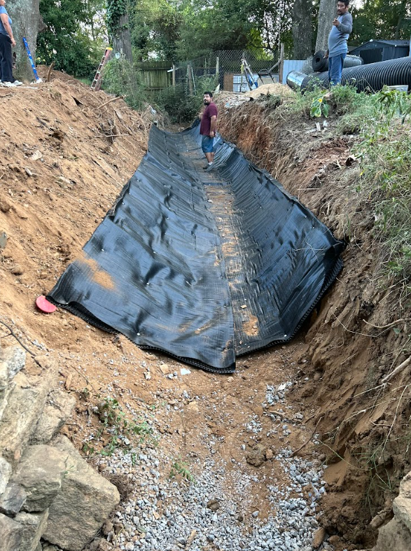 Crew lining a wide excavated trench with fabric and gravel before backfilling