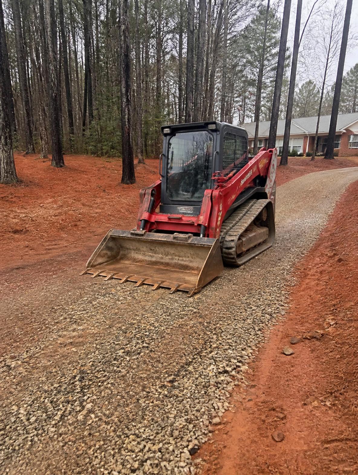 Compact track loader with a tooth bucket working a gravel driveway in a wooded Georgia yard