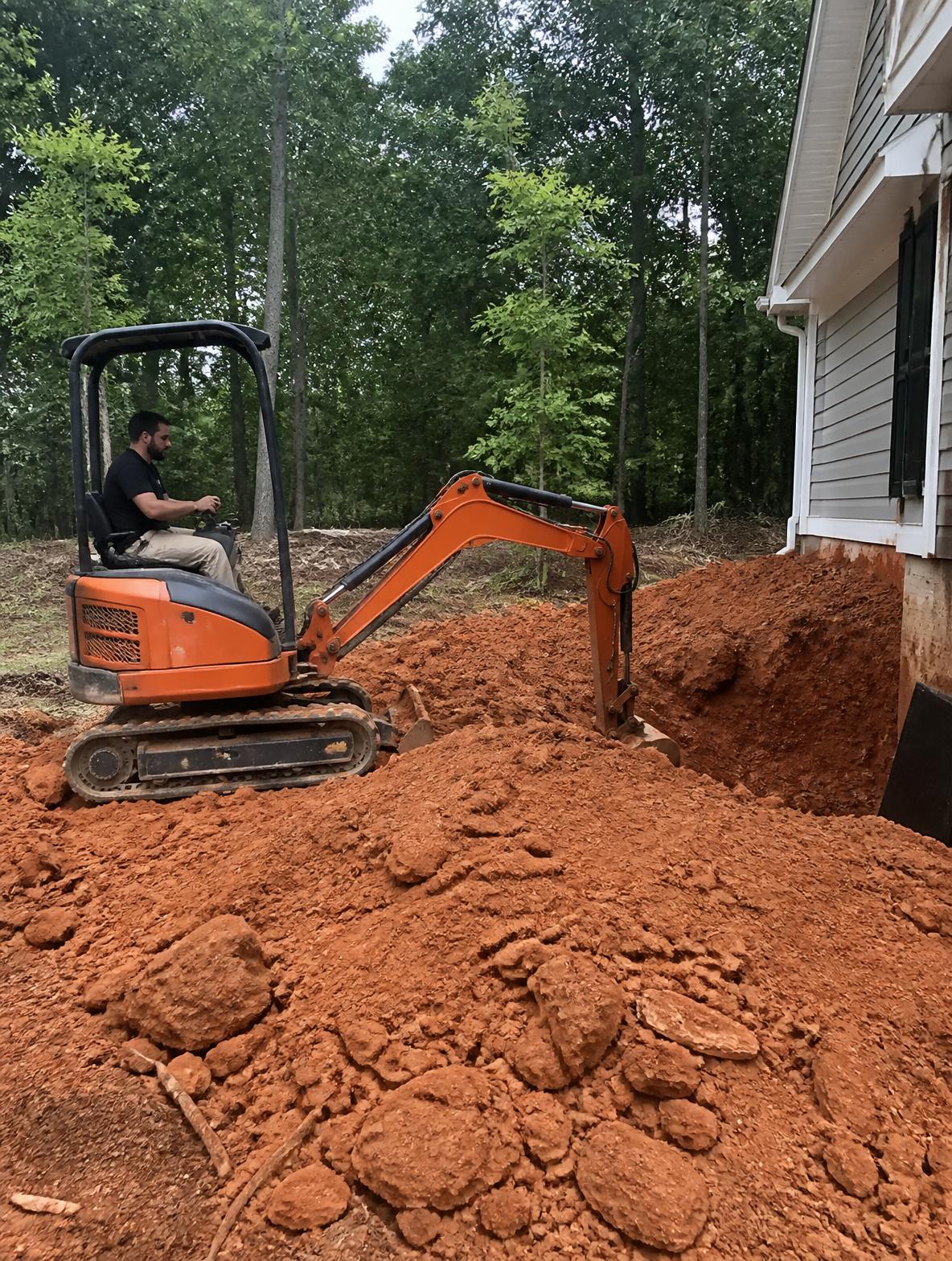 Mini excavator digging red clay soil beside a home foundation